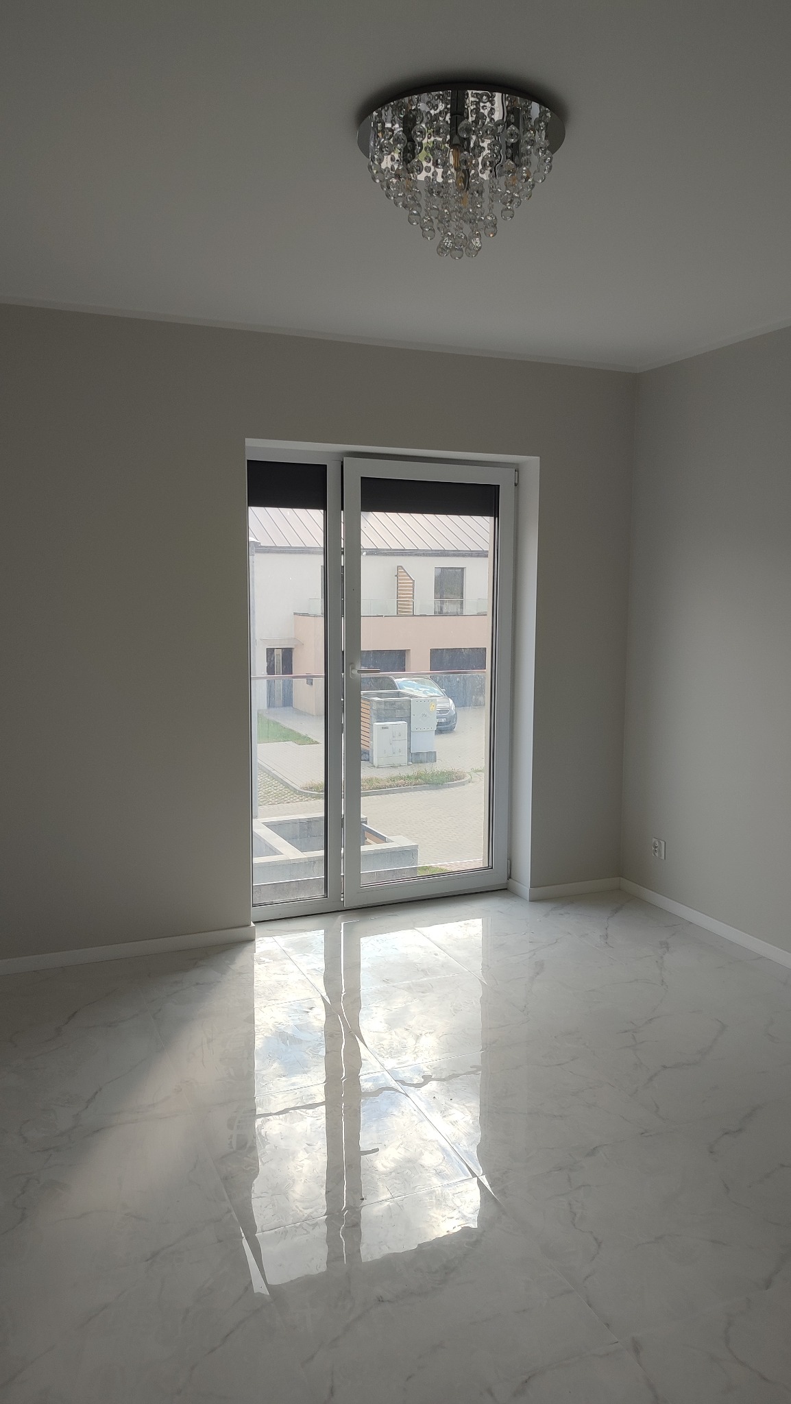 Pusty pokój z błyszczącą marmurową podłogą, nowoczesnym kryształowym żyrandolem i dużym oknem przesuwnym z widokiem na dom i zaparkowany samochód.