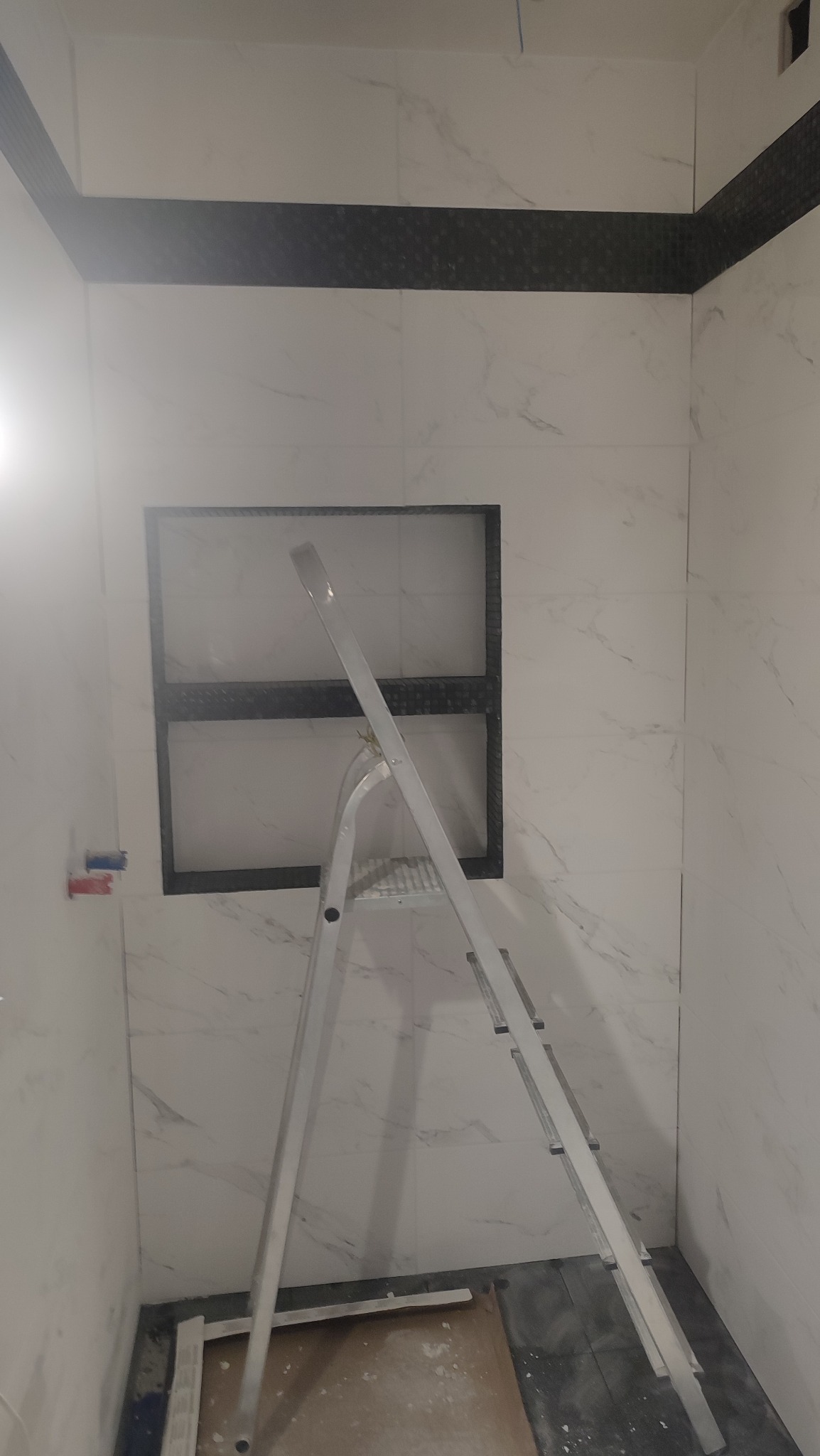 Wnętrze łazienki w trakcie wykańczania, widoczne białe płytki imitujące marmur, czarna listwa dekoracyjna, wnęka w ścianie z czarnym obramowaniem oraz aluminiowa drabina.