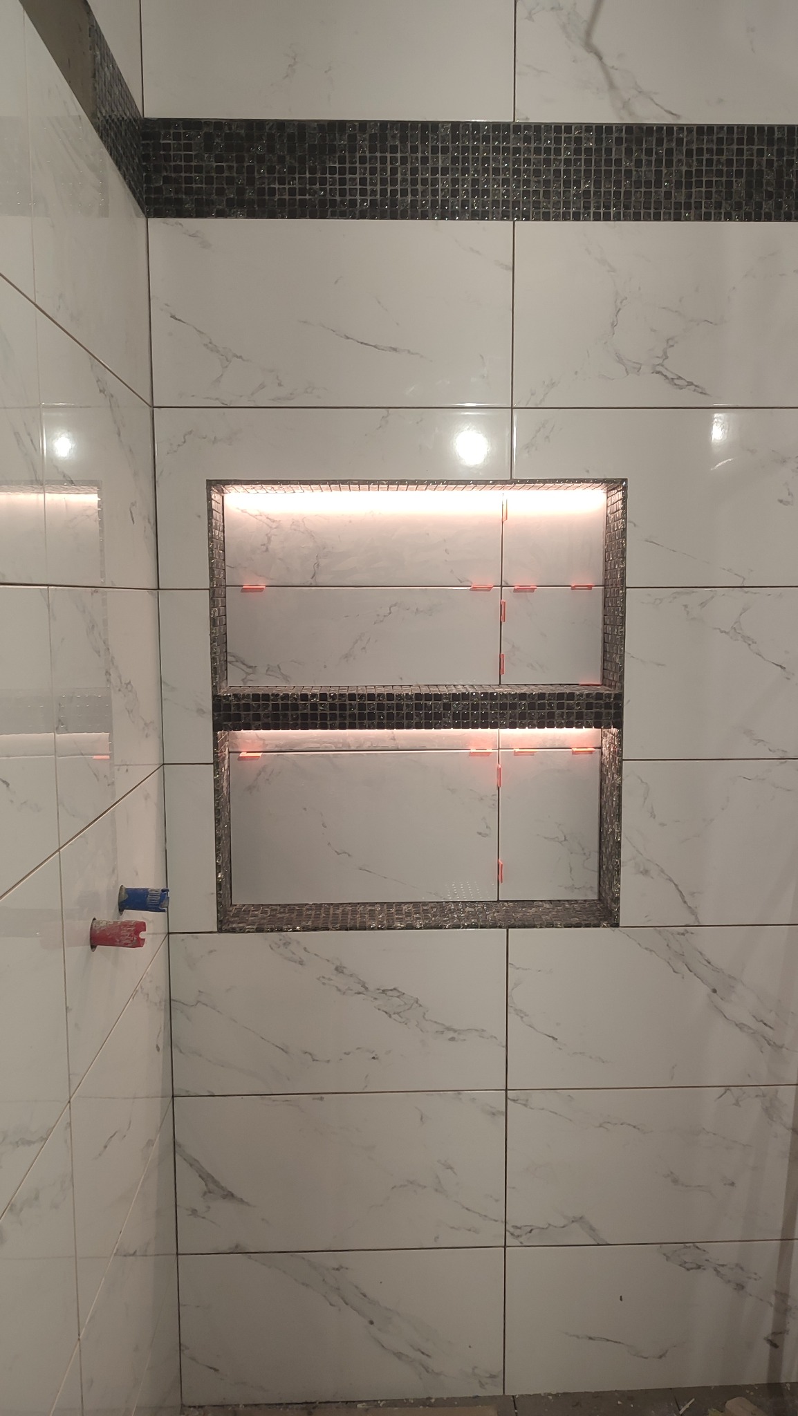 Wnęka z podświetleniem LED w ścianie wyłożonej białymi płytkami imitującymi marmur, z dekoracyjnym pasem mozaiki na górze, w trakcie montażu płytek, widoczne krzyżyki dystansowe.