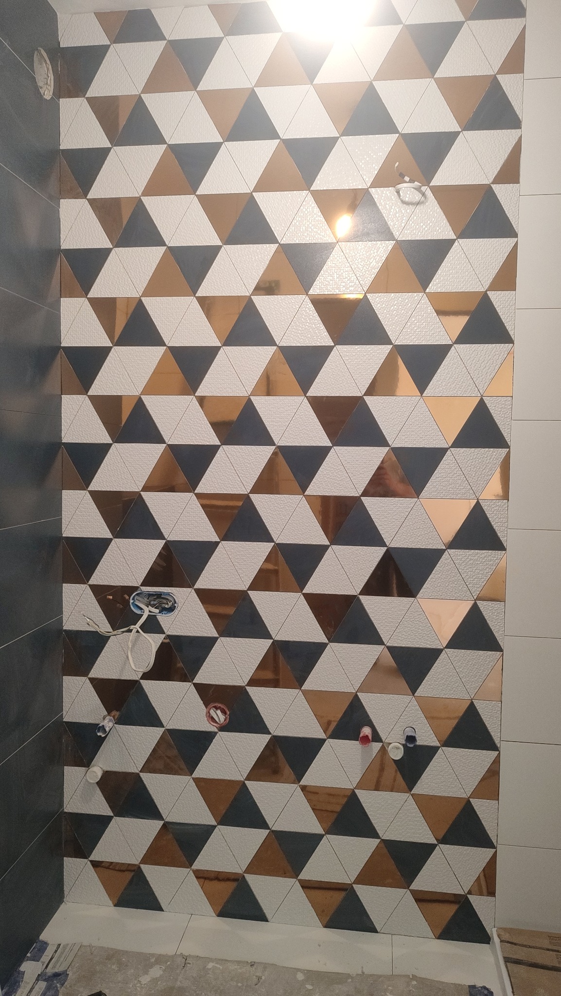Ściana łazienki wyłożona trójkątnymi płytkami w kolorach białym, szarym i miedzianym. Widoczne wystające rury instalacji wodnej i elektrycznej przed montażem armatury.