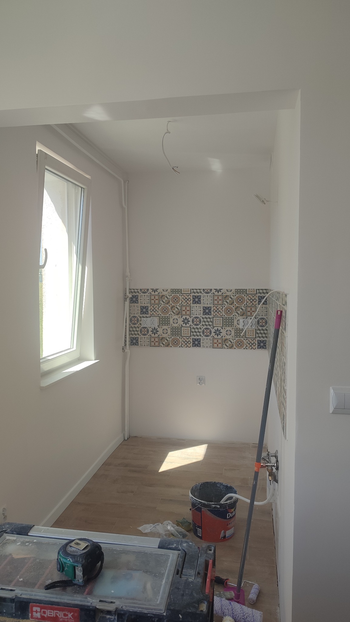 Wnętrze łazienki w trakcie remontu: widoczne płytki dekoracyjne na ścianie, wiadro z farbą Dulux, narzędzia glazurnicze i fragment okna.