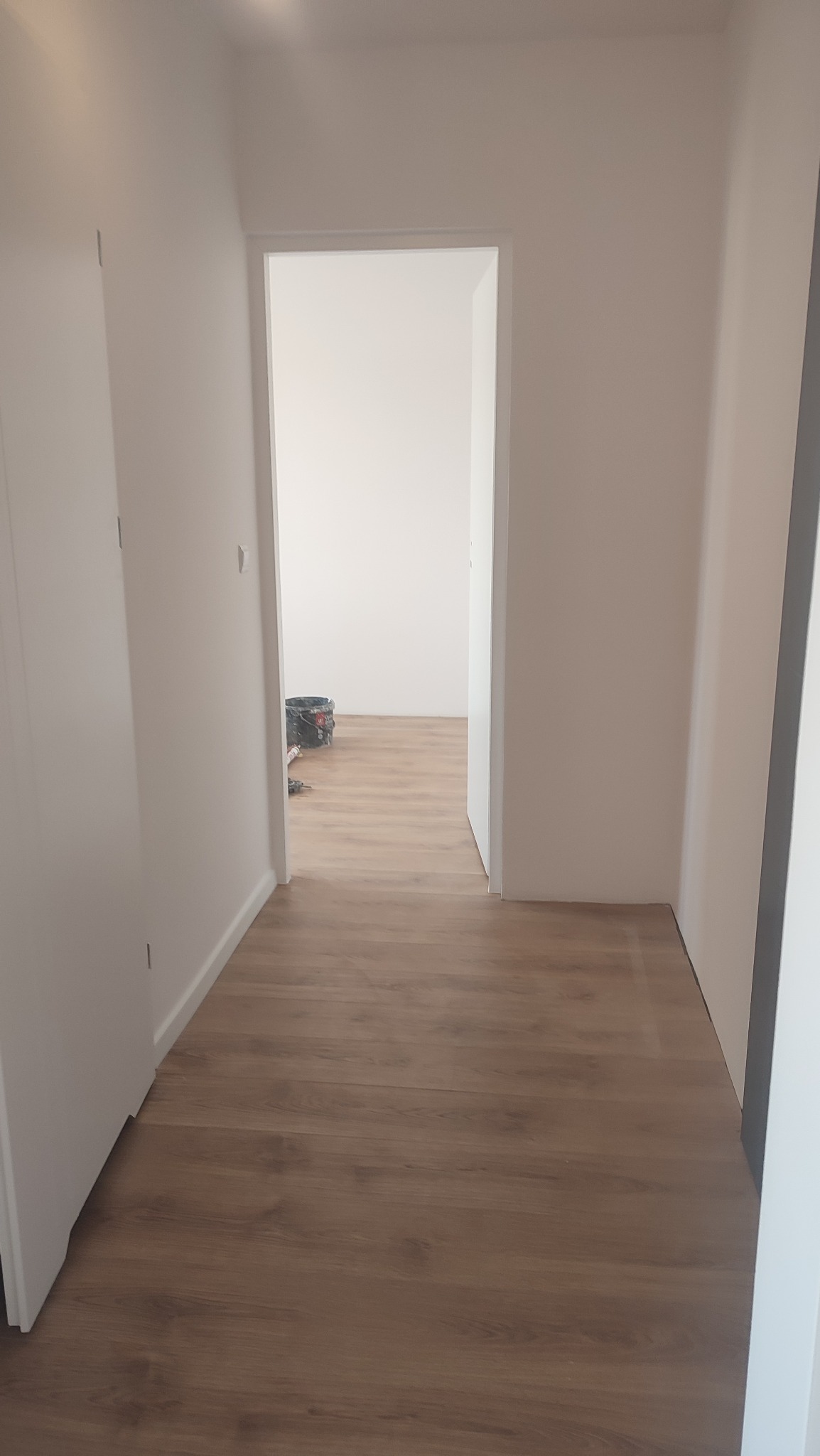 Korytarz z nową podłogą laminowaną o ciepłym odcieniu drewna, świeżo pomalowane białe ściany i widok na sąsiednie pomieszczenie z wiadrem i narzędziami malarskimi w tle.