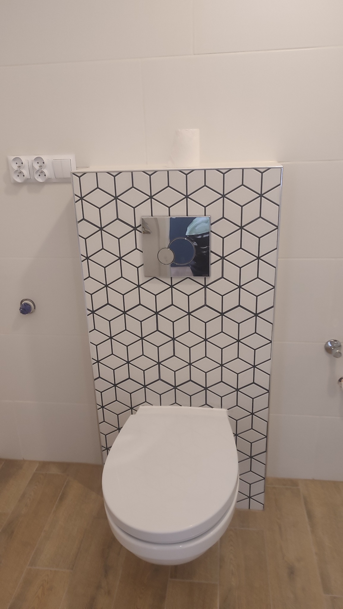 Wisząca toaleta z geometryczną mozaiką na zabudowie stelaża, widoczne gniazdka elektryczne i rolka papieru toaletowego.