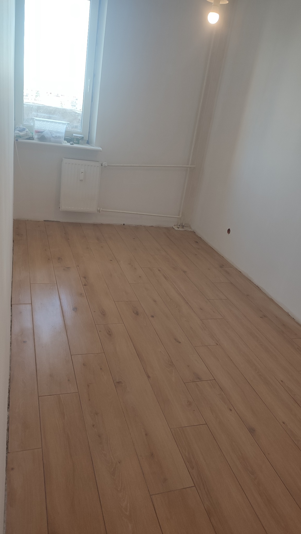 Jasne panele podłogowe ułożone w pustym pokoju z białymi ścianami, grzejnikiem pod oknem i pojedynczą żarówką na suficie.