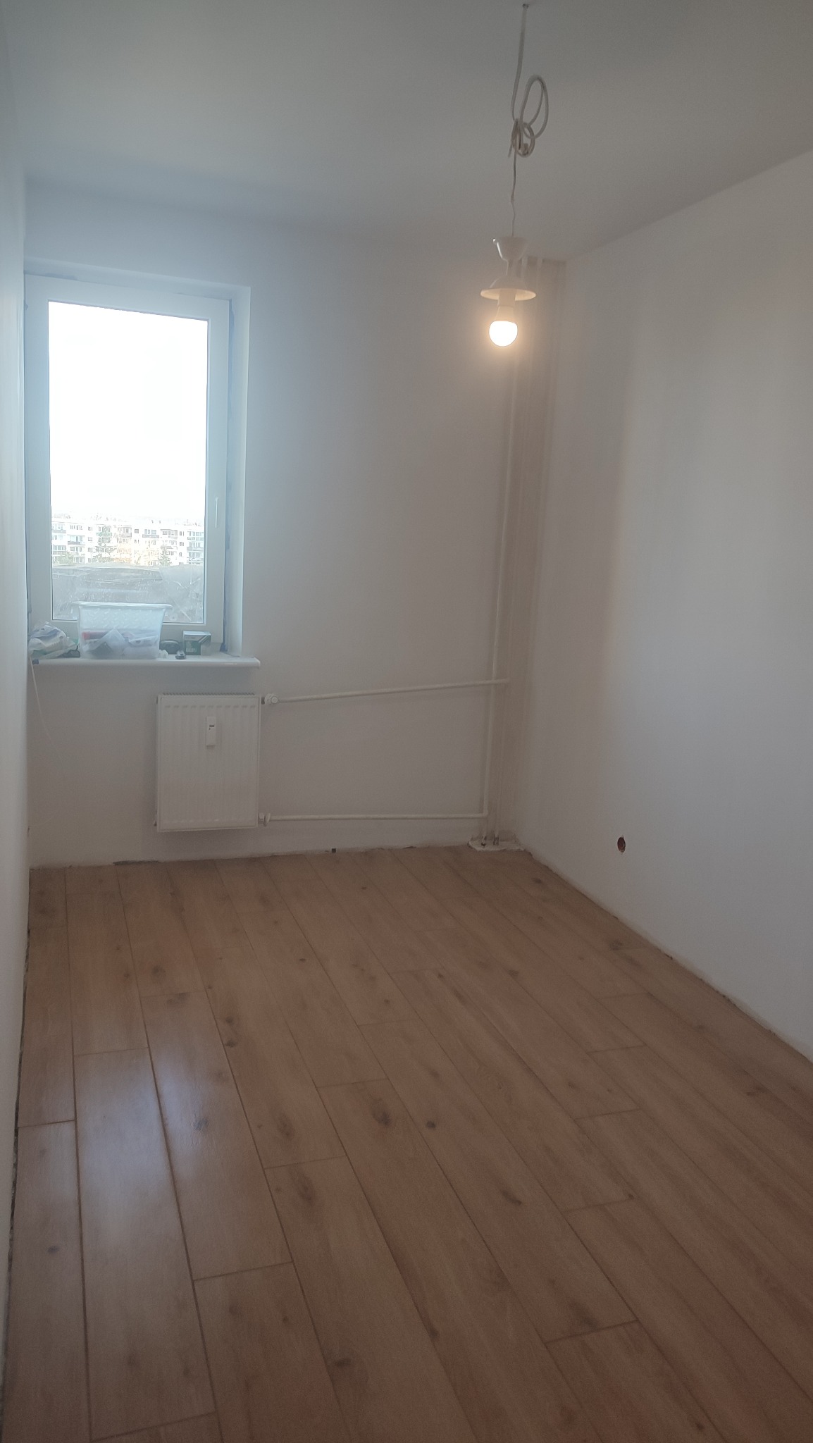 Pusty pokój z nową podłogą z paneli, grzejnikiem pod oknem i wiszącą żarówką. Widok na budynki za oknem.