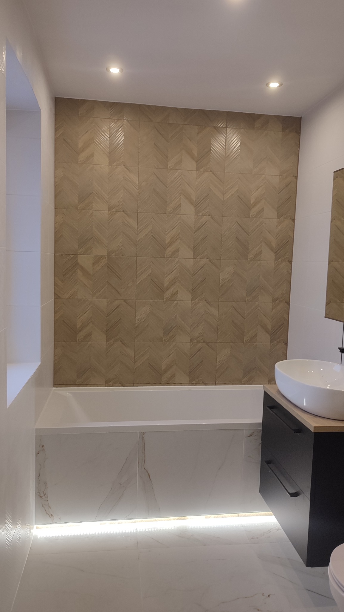 Wykończona łazienka z drewnopodobną ścianą w jodełkę, białą wanną i podświetleniem LED pod wanną, minimalistyczna umywalka nablatowa na czarnej szafce.