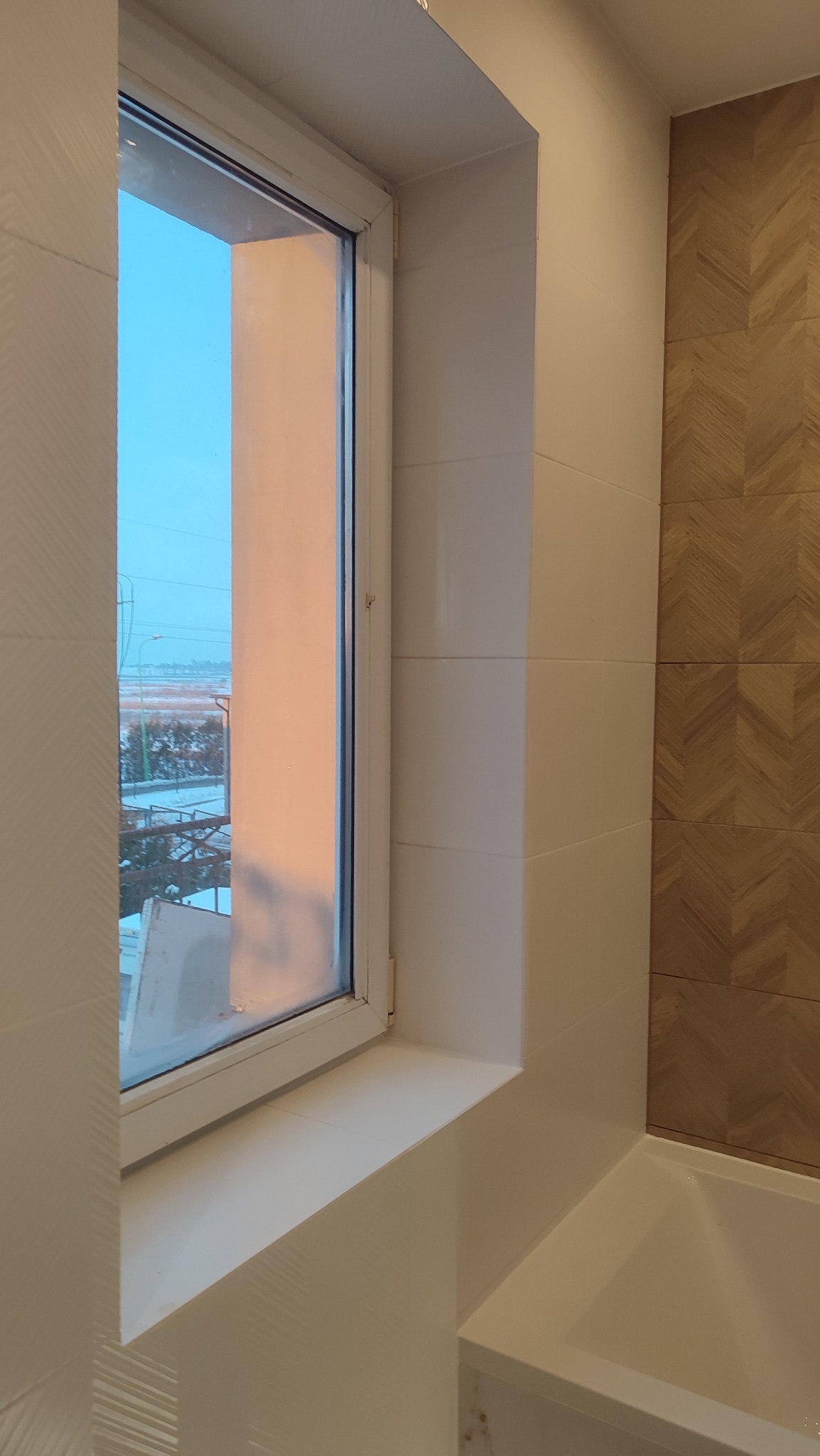 Okno w łazience z widokiem na zimowy krajobraz, białe płytki i dekoracyjna ściana z motywem jodełki w odcieniach brązu, fragment wanny widoczny w rogu.