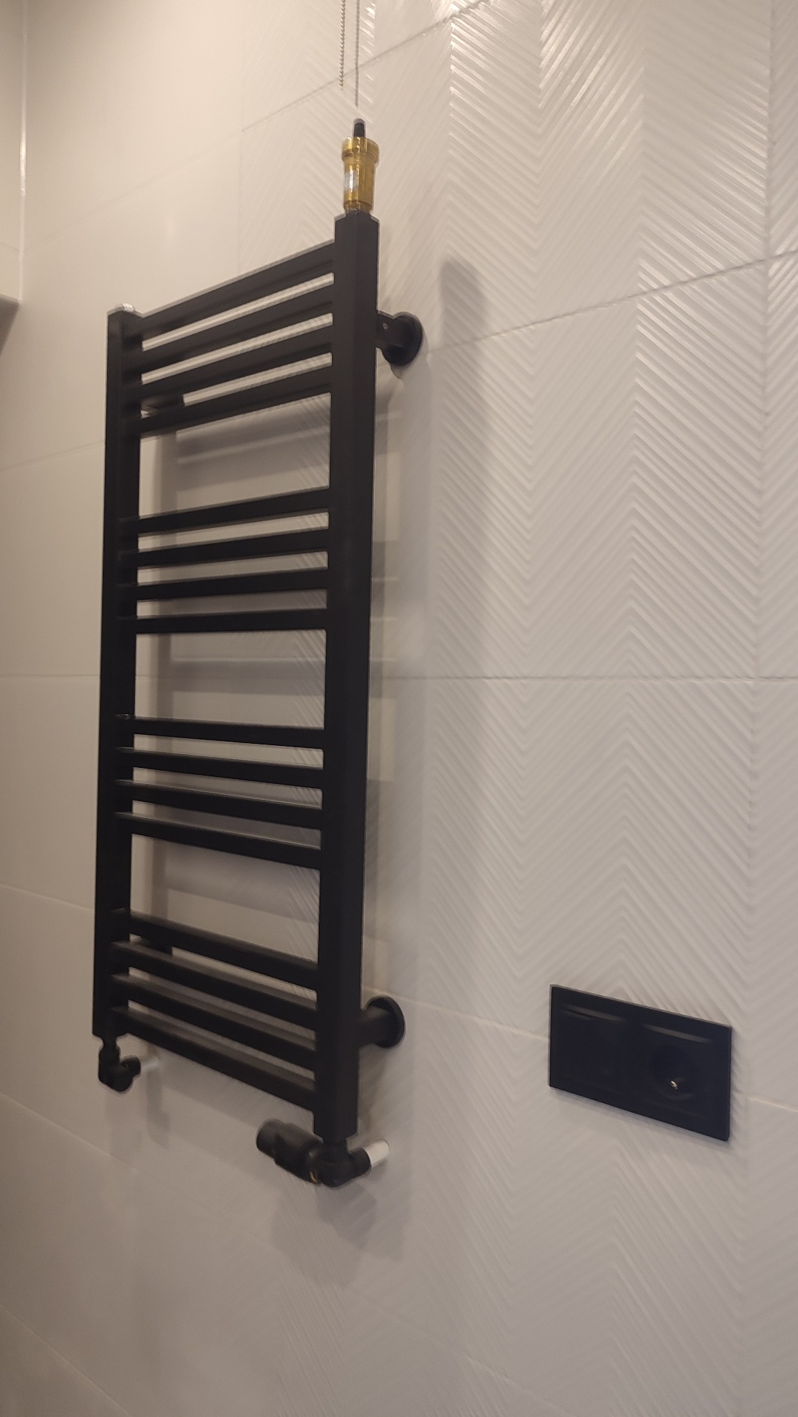 Czarny grzejnik łazienkowy drabinkowy zamontowany na białej ścianie z subtelnym wzorem chevron, widoczny zawór termostatyczny i czarna maskownica przyłącza.