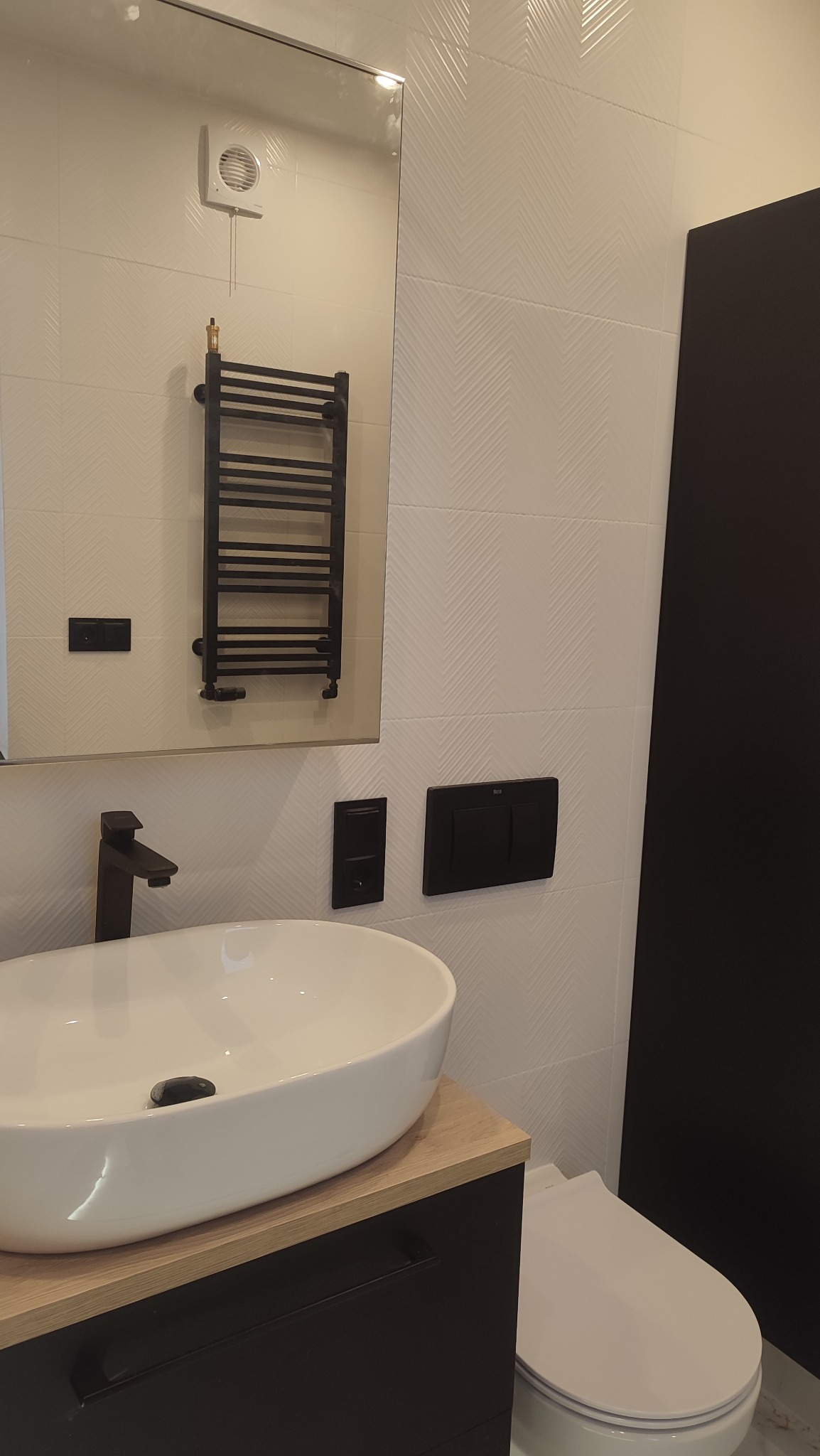 Nowoczesna łazienka z owalną białą umywalką na drewnopodobnym blacie, czarną armaturą, grzejnikiem drabinkowym i toaletą; białe płytki w jodełkę na ścianie.