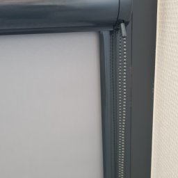 ROLOTEKA - marka należąca do VERSAPOINT SPÓŁKA Z OGRANICZONĄ ODPOWIEDZIALNOŚCIĄ - Szara roleta wewnętrzna zwinięta przy oknie, widoczny mechanizm łańcuszkowy. Ściana w tle o delikatnej strukturze.