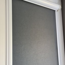 ROLOTEKA - marka należąca do VERSAPOINT SPÓŁKA Z OGRANICZONĄ ODPOWIEDZIALNOŚCIĄ - Szara, zwinięta zasłona okienna w białej ramie, z widocznym mechanizmem łańcuszkowym po prawej stronie.