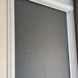 Szara, zwinięta zasłona okienna w białej ramie, z widocznym mechanizmem łańcuszkowym po prawej stronie.