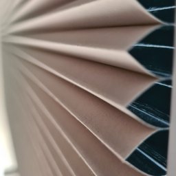 ROLOTEKA - marka należąca do VERSAPOINT SPÓŁKA Z OGRANICZONĄ ODPOWIEDZIALNOŚCIĄ - Beżowe, plisowane osłony okienne w zbliżeniu, widoczne delikatne załamania i struktura materiału na tle ciemnego okna.