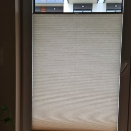 ROLOTEKA - marka należąca do VERSAPOINT SPÓŁKA Z OGRANICZONĄ ODPOWIEDZIALNOŚCIĄ - Beżowa, plisowana osłona okienna, częściowo zasłonięta, widok na balkon z szarymi balustradami i sztuczną trawą.