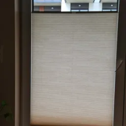 Beżowa, plisowana osłona okienna, częściowo zasłonięta, widok na balkon z szarymi balustradami i sztuczną trawą.