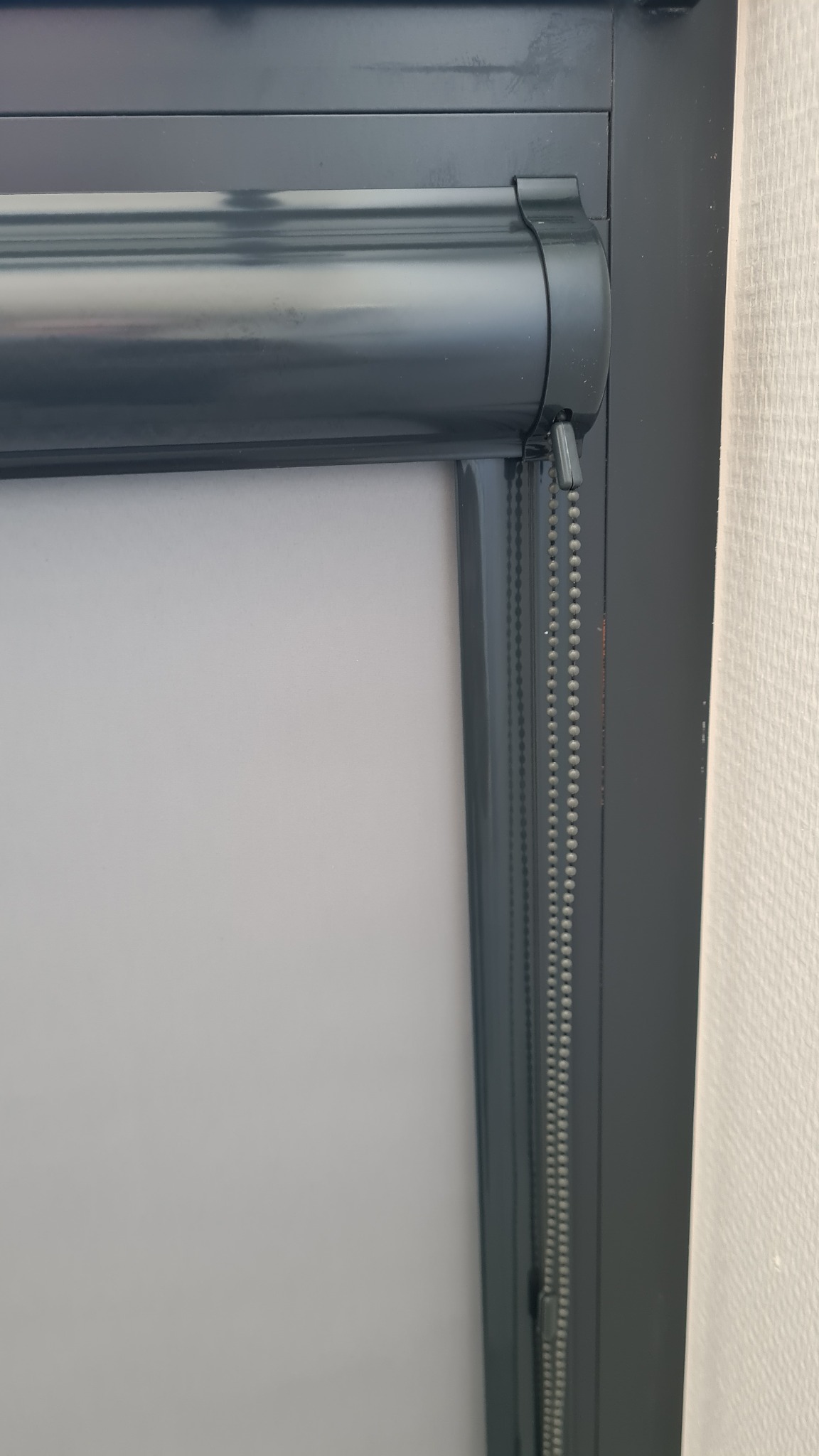 Szara roleta wewnętrzna zwinięta przy oknie, widoczny mechanizm łańcuszkowy. Ściana w tle o delikatnej strukturze.