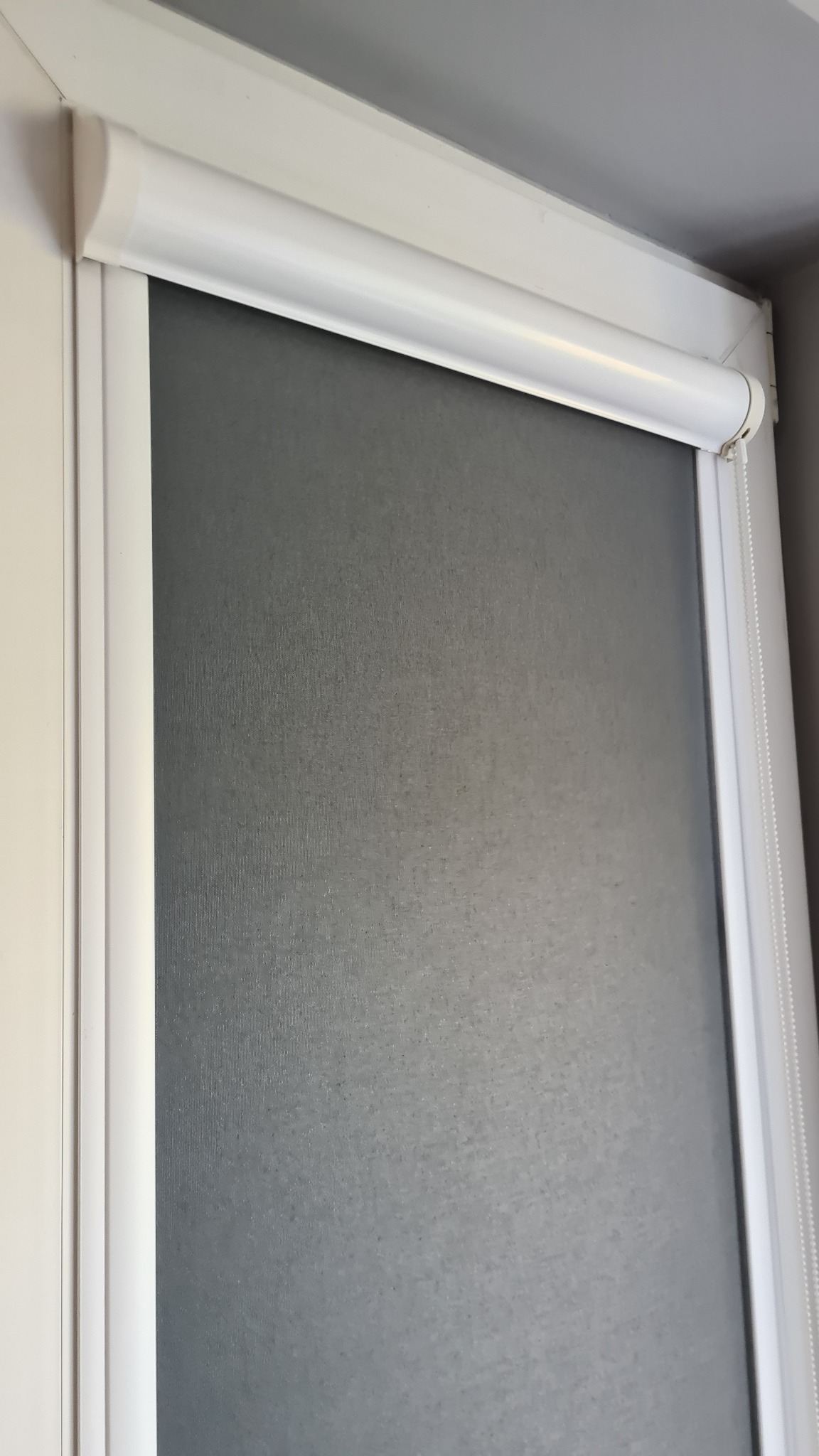 Szara, zwinięta zasłona okienna w białej ramie, z widocznym mechanizmem łańcuszkowym po prawej stronie.