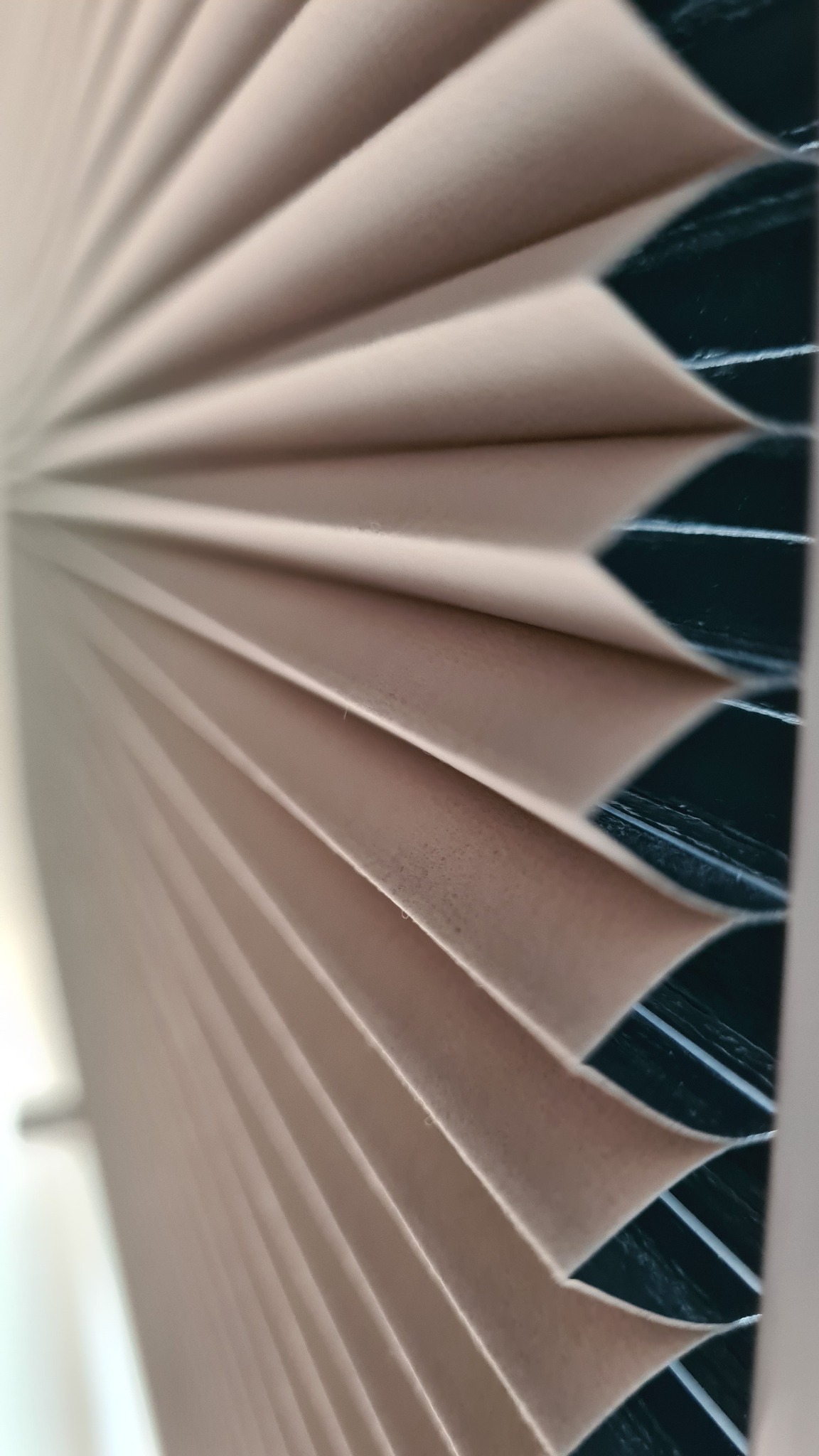 Beżowe, plisowane osłony okienne w zbliżeniu, widoczne delikatne załamania i struktura materiału na tle ciemnego okna.