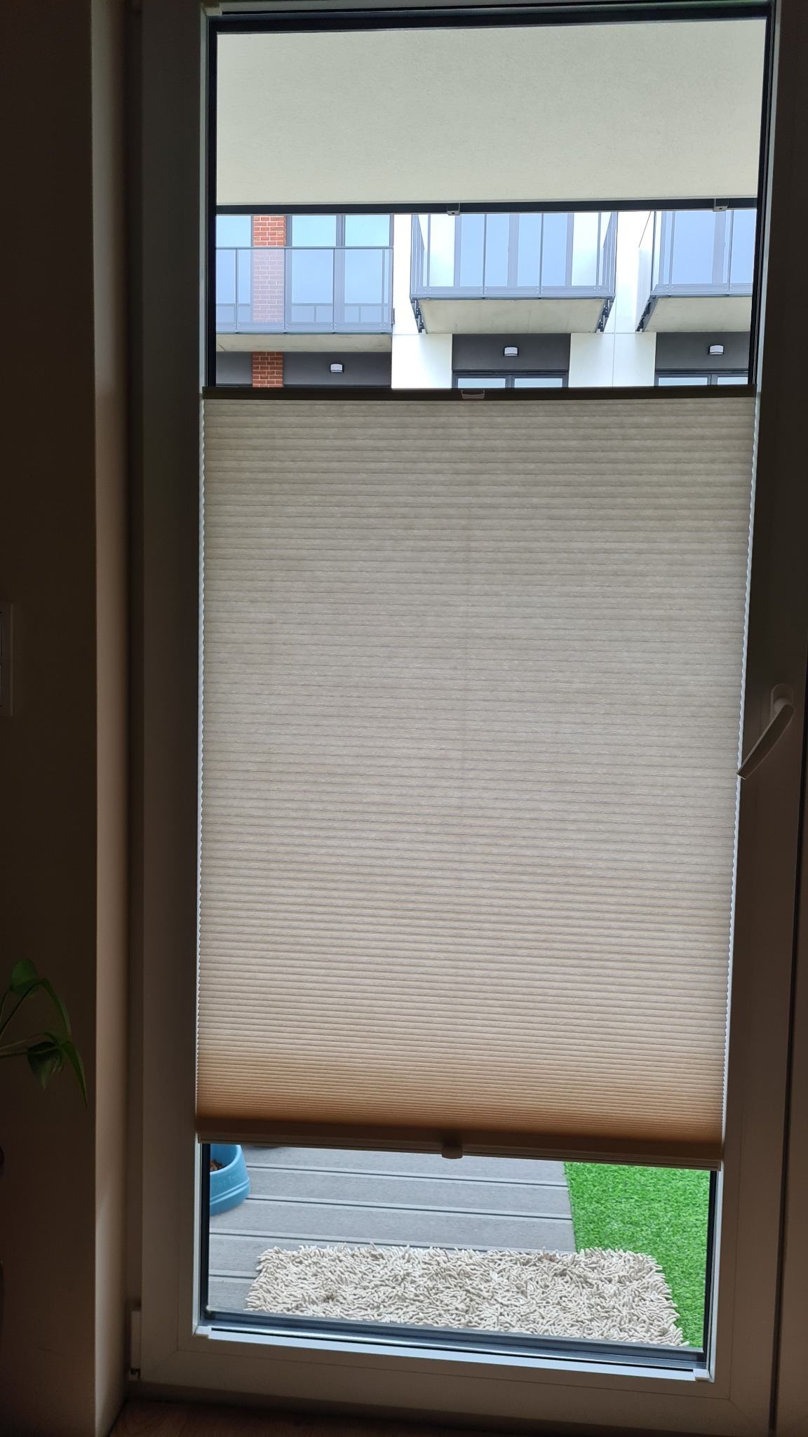 Beżowa, plisowana osłona okienna, częściowo zasłonięta, widok na balkon z szarymi balustradami i sztuczną trawą.