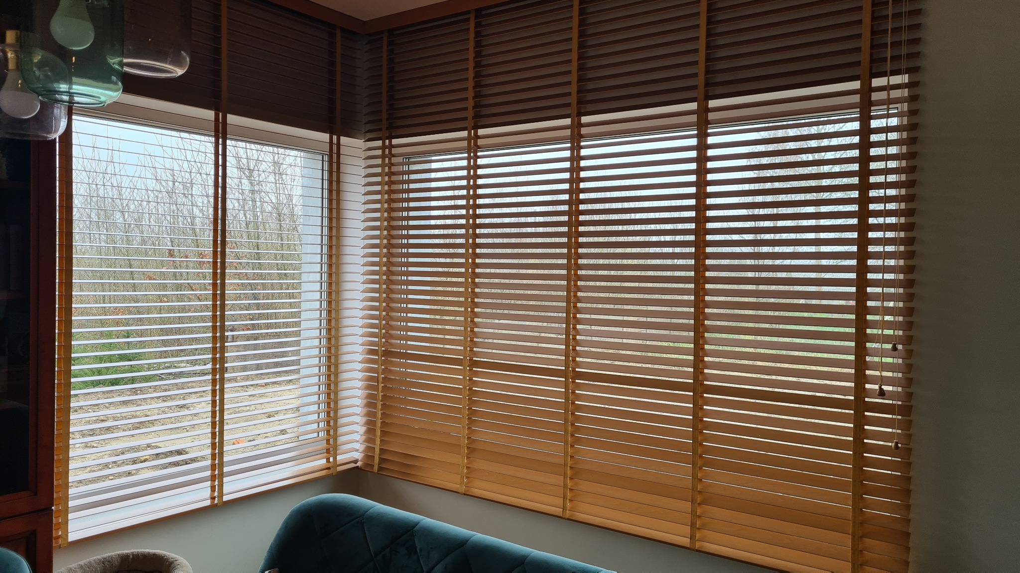 Okna z drewnianymi żaluzjami w kolorze miodowym, częściowo zasłonięte, widok na drzewa za oknem, fragment zielonego welurowego mebla w dolnej części kadru.