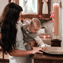 Piotr Wojnarowski - Matka trzymająca dziecko nad zdobioną misą podczas chrztu w kościele, w tle ołtarz i świeca paschalna. Ujęcie z bliska, ciepłe światło.