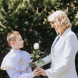 Piotr Wojnarowski - Chłopiec w stroju komunijnym i kobieta w białym żakiecie wymieniają się białą różą na tle zieleni. Jasne, naturalne światło, portret z uroczystości.