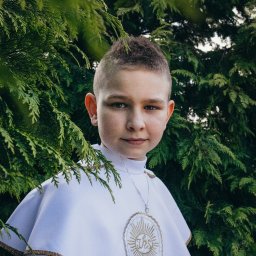 Piotr Wojnarowski - Portret chłopca w stroju komunijnym, z medalikiem, na tle zieleni. Ujęcie z bliska, z naturalnym oświetleniem i delikatnym uśmiechem.
