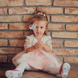 Piotr Wojnarowski - Dziewczynka w różowej sukience siedzi na podłodze z ceglaną ścianą w tle, trzymając w ręku srebrną różdżkę w kształcie serca. Na podłodze leży korona.