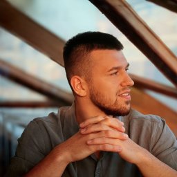 Piotr Wojnarowski - Młody mężczyzna z brodą, w szarej koszuli, spogląda w bok z zamyśleniem na tle rozmytej konstrukcji. Dłonie splecione przed sobą. Ujęcie z profilu.