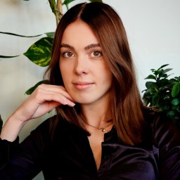 Piotr Wojnarowski - Portret kobiety z brązowymi włosami, w czarnej satynowej koszuli, z dłonią pod brodą, na tle zielonych roślin. Subtelny makijaż i złoty naszyjnik.