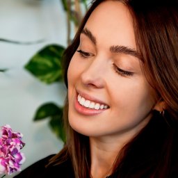 Piotr Wojnarowski - Uśmiechnięta brunetka z kwiatem w dłoni, spoglądająca w dół. Naturalne światło, delikatny makijaż, zieleń w tle. Zdjęcie profilowe, portret.