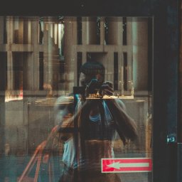 Piotr Wojnarowski - Mężczyzna robi zdjęcie aparatem w odbiciu szyby. Widoczne miejskie tło i neonowy znak. Ujęcie z bliska, z efektem nałożonych warstw.