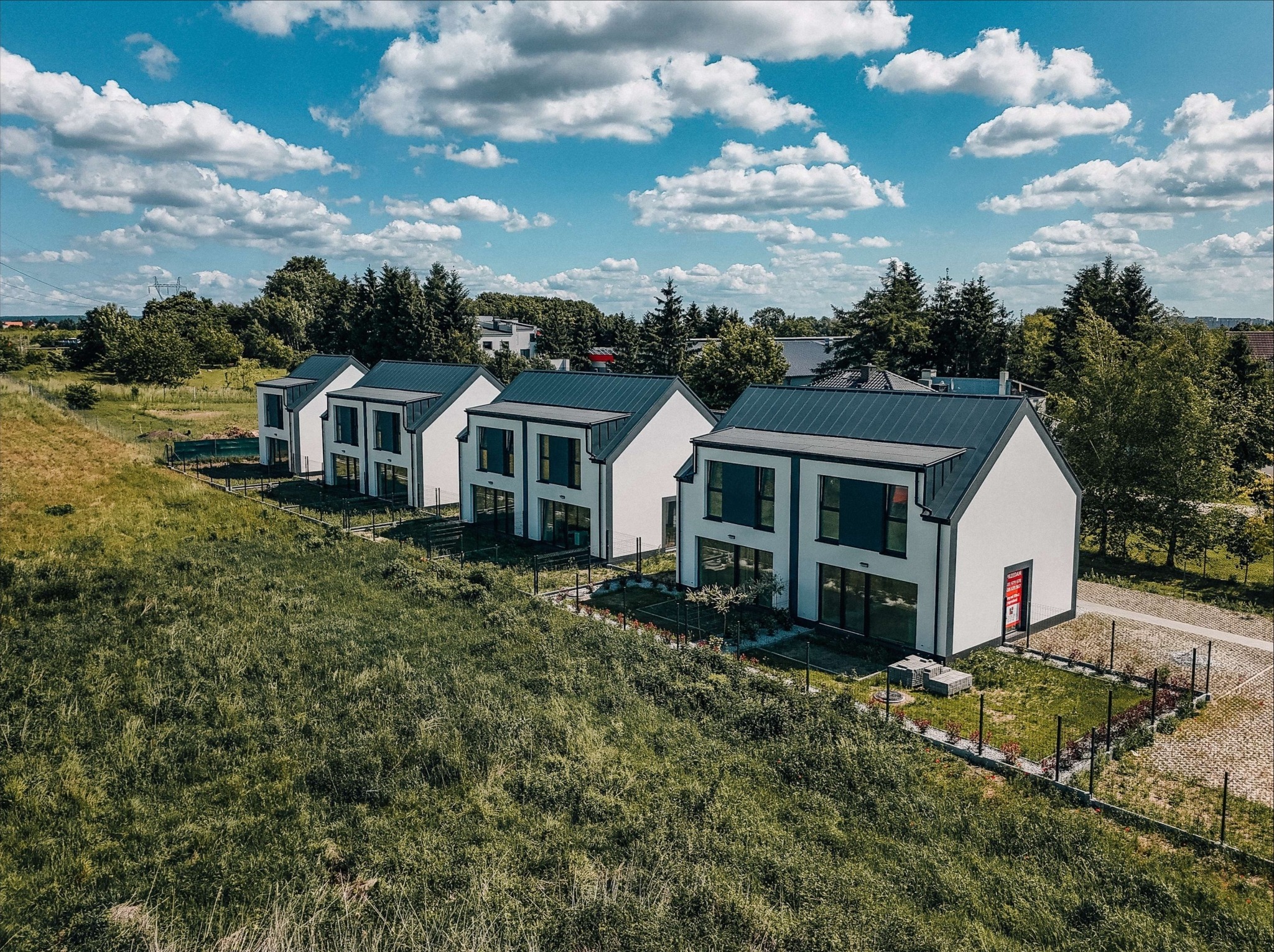 Nowoczesne, szeregowe domy jednorodzinne z grafitowymi dachami i białymi elewacjami, otoczone zielenią, sfotografowane z lotu ptaka w słoneczny dzień.