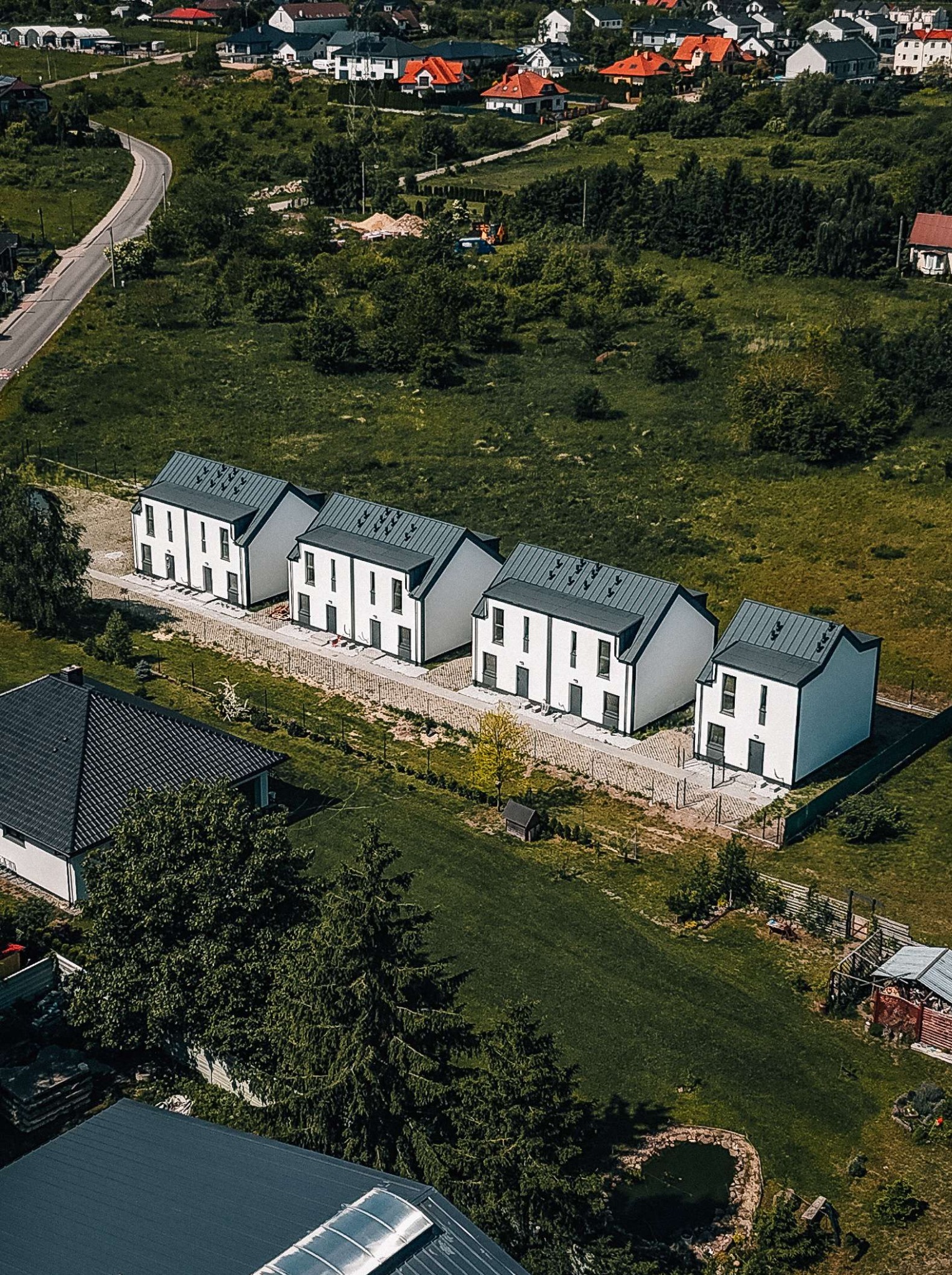 Widok z lotu ptaka na szereg identycznych, nowoczesnych domów z ciemnymi dachami i białymi ścianami. Otoczenie to zielone tereny z pojedynczymi drzewami i drogą w tle.