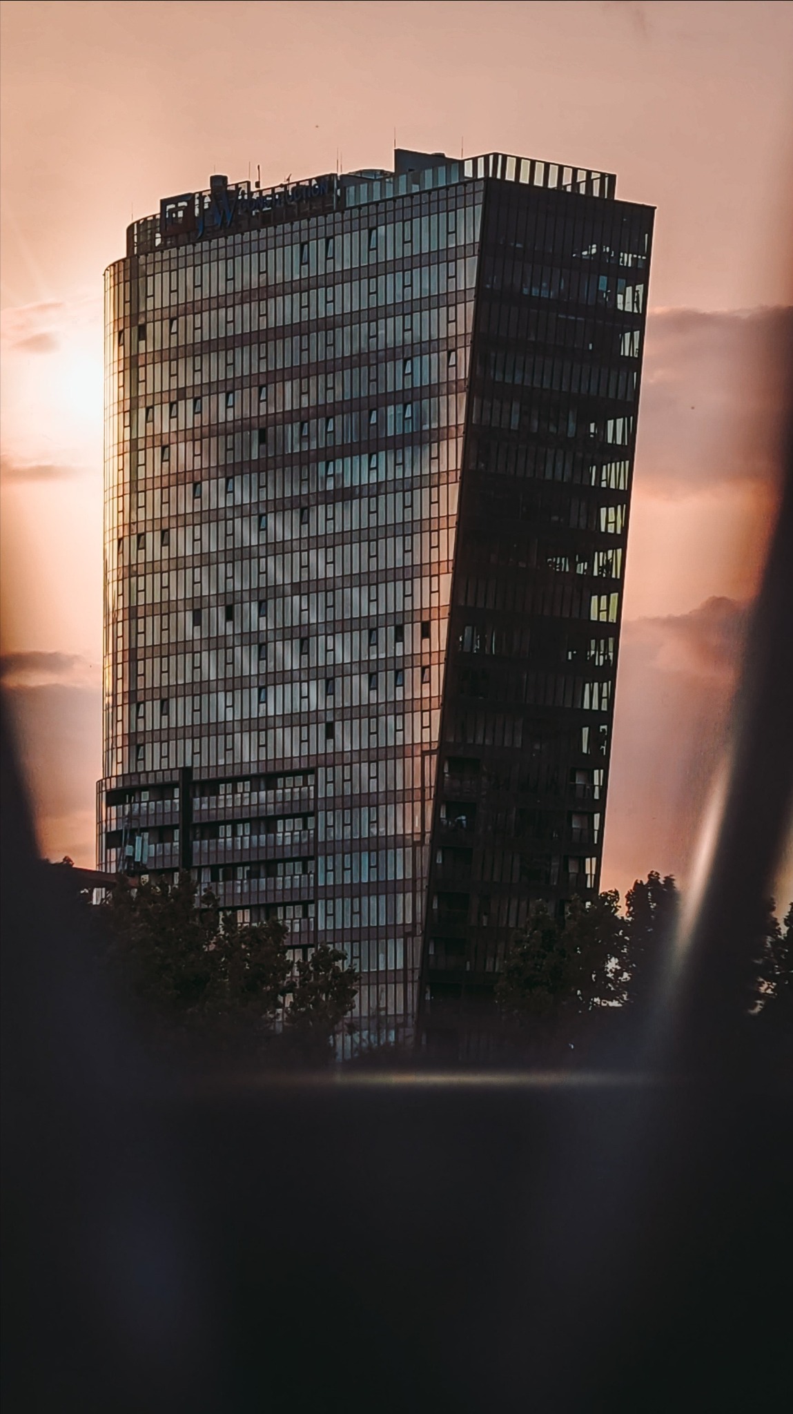 Nowoczesny, wysoki budynek biurowy z licznymi oknami i asymetryczną bryłą na tle zachodzącego słońca. Ujęcie z dołu, z widocznymi drzewami w dolnej części kadru.
