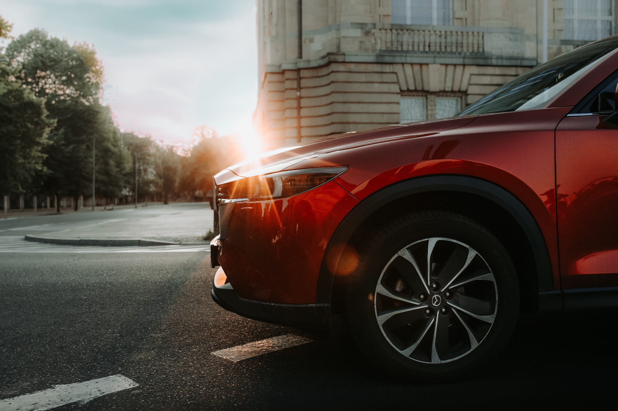 Czerwona Mazda CX-5 na tle miejskiej architektury, oświetlona promieniami zachodzącego słońca. Widoczny fragment jezdni i budynek z elementami klasycystycznymi.