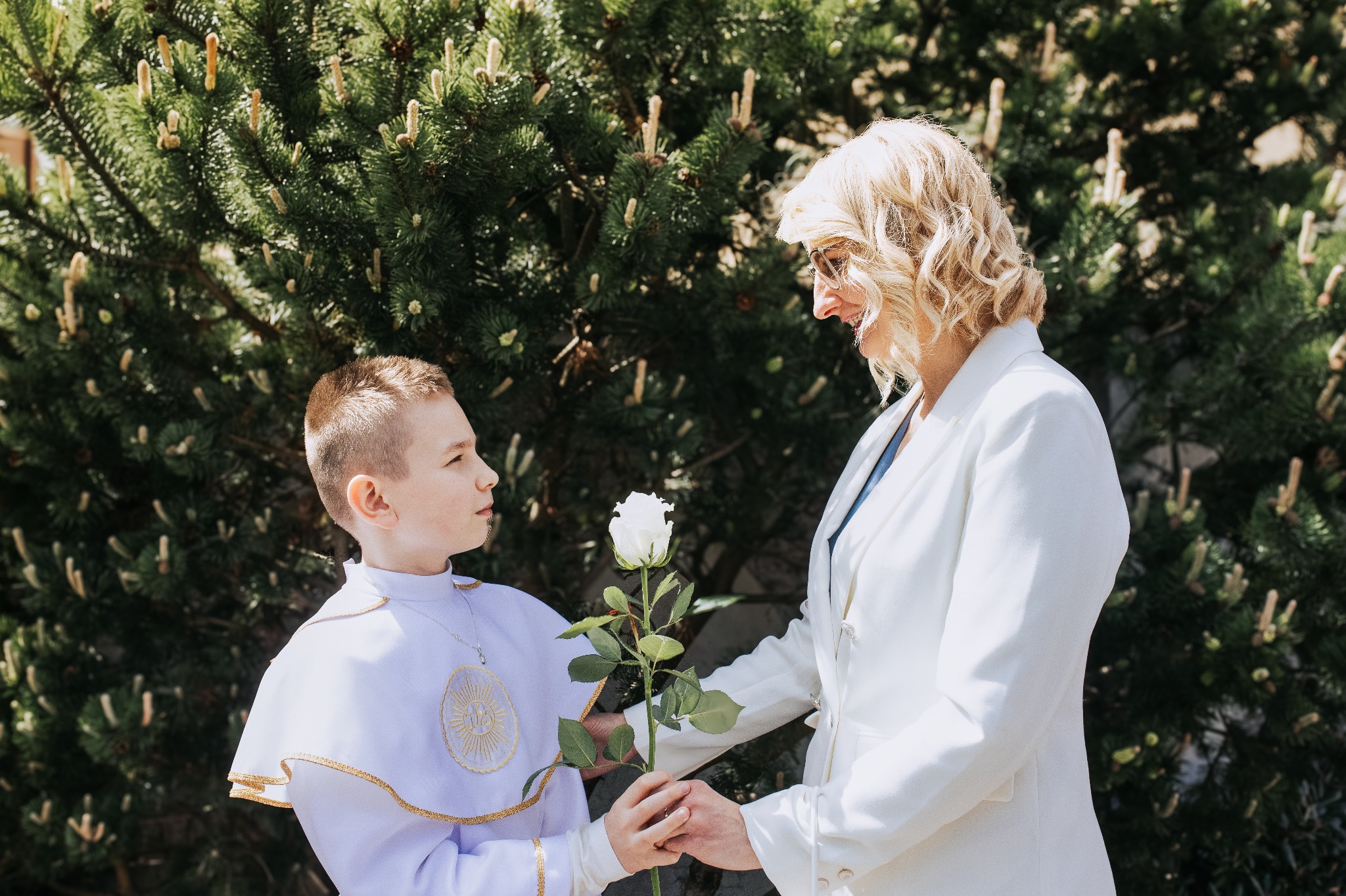 Chłopiec w stroju komunijnym i kobieta w białym żakiecie wymieniają się białą różą na tle zieleni. Jasne, naturalne światło, portret z uroczystości.