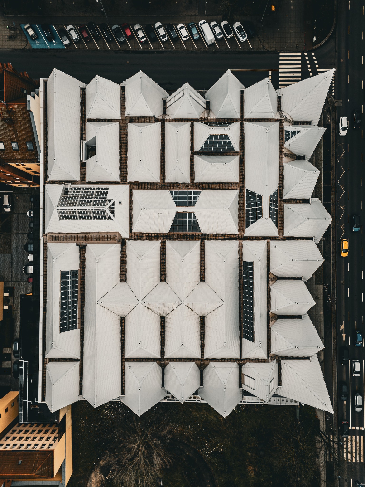 Geometryczny dach budynku z blachy, widziany z lotu ptaka. Regularny wzór z trójkątnych i prostokątnych elementów, szara kolorystyka, z elementami zieleni i ulicą z samochodami.