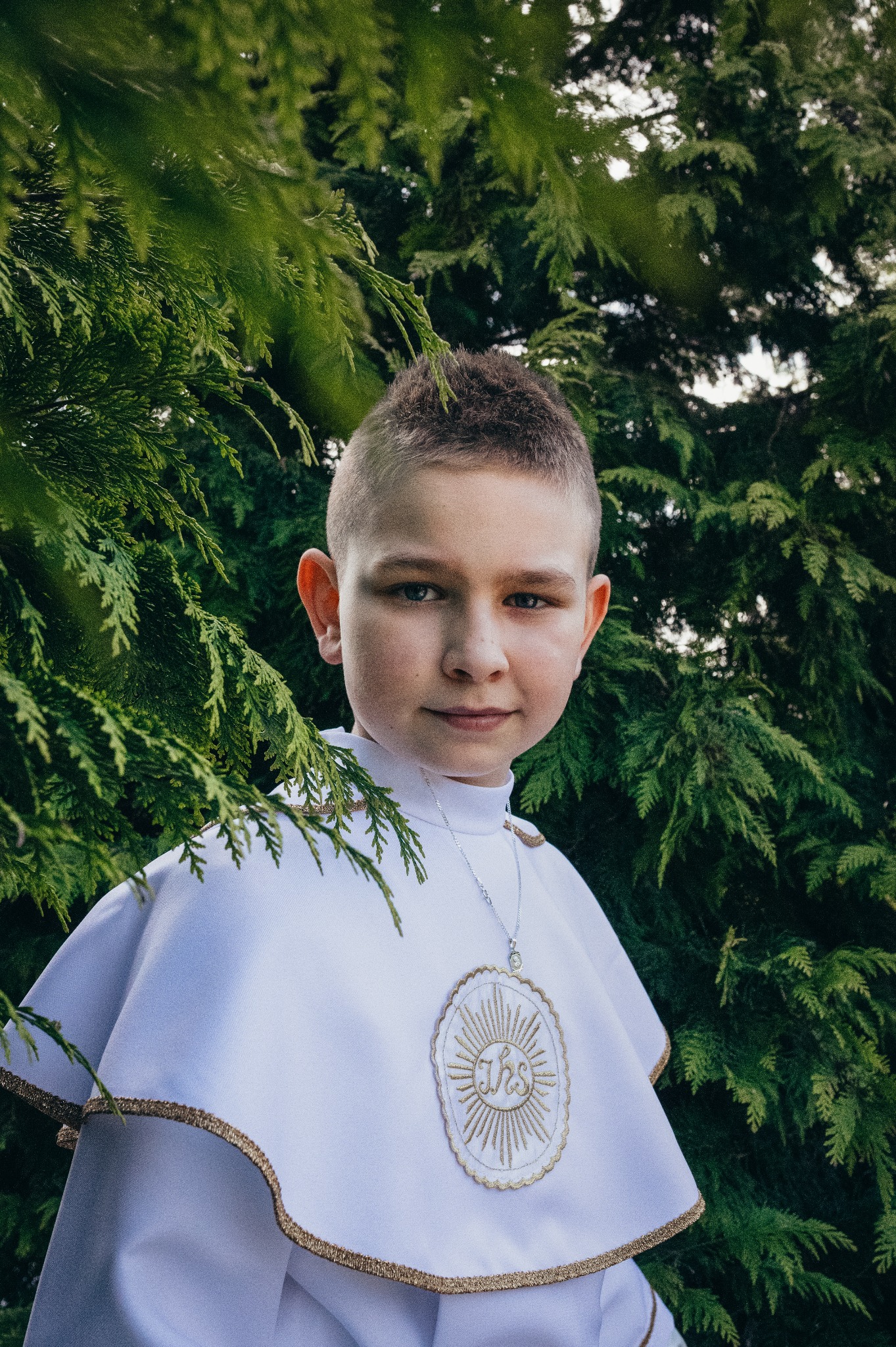 Portret chłopca w stroju komunijnym, z medalikiem, na tle zieleni. Ujęcie z bliska, z naturalnym oświetleniem i delikatnym uśmiechem.