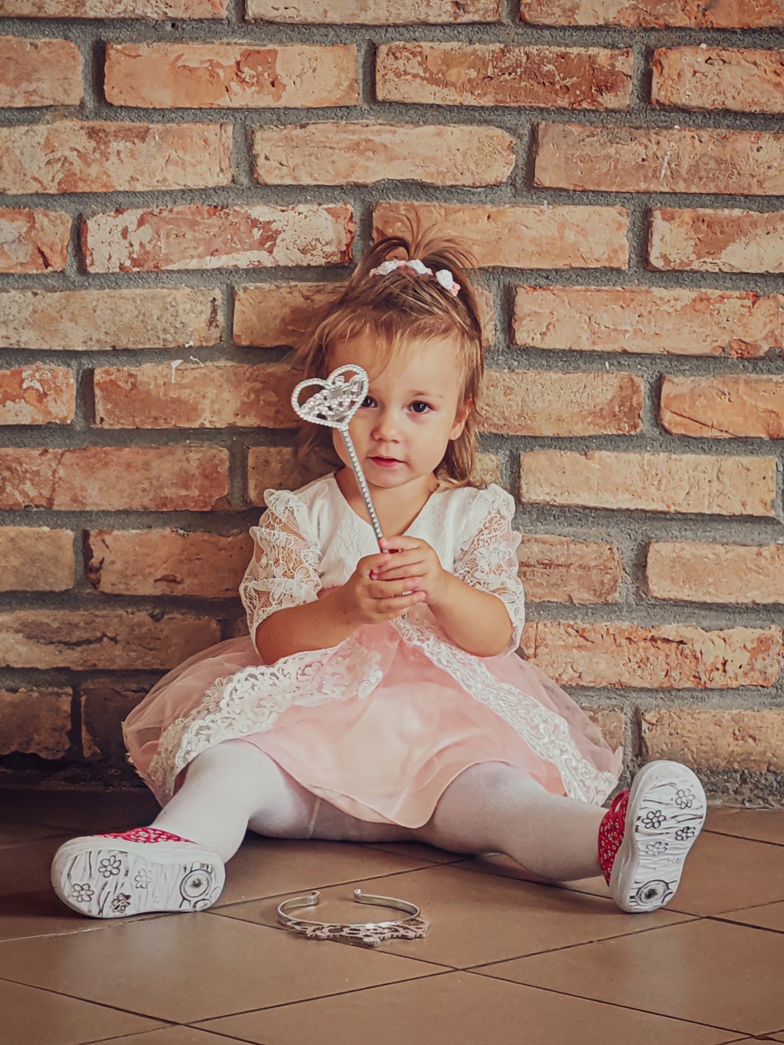 Dziewczynka w różowej sukience siedzi na podłodze z ceglaną ścianą w tle, trzymając w ręku srebrną różdżkę w kształcie serca. Na podłodze leży korona.