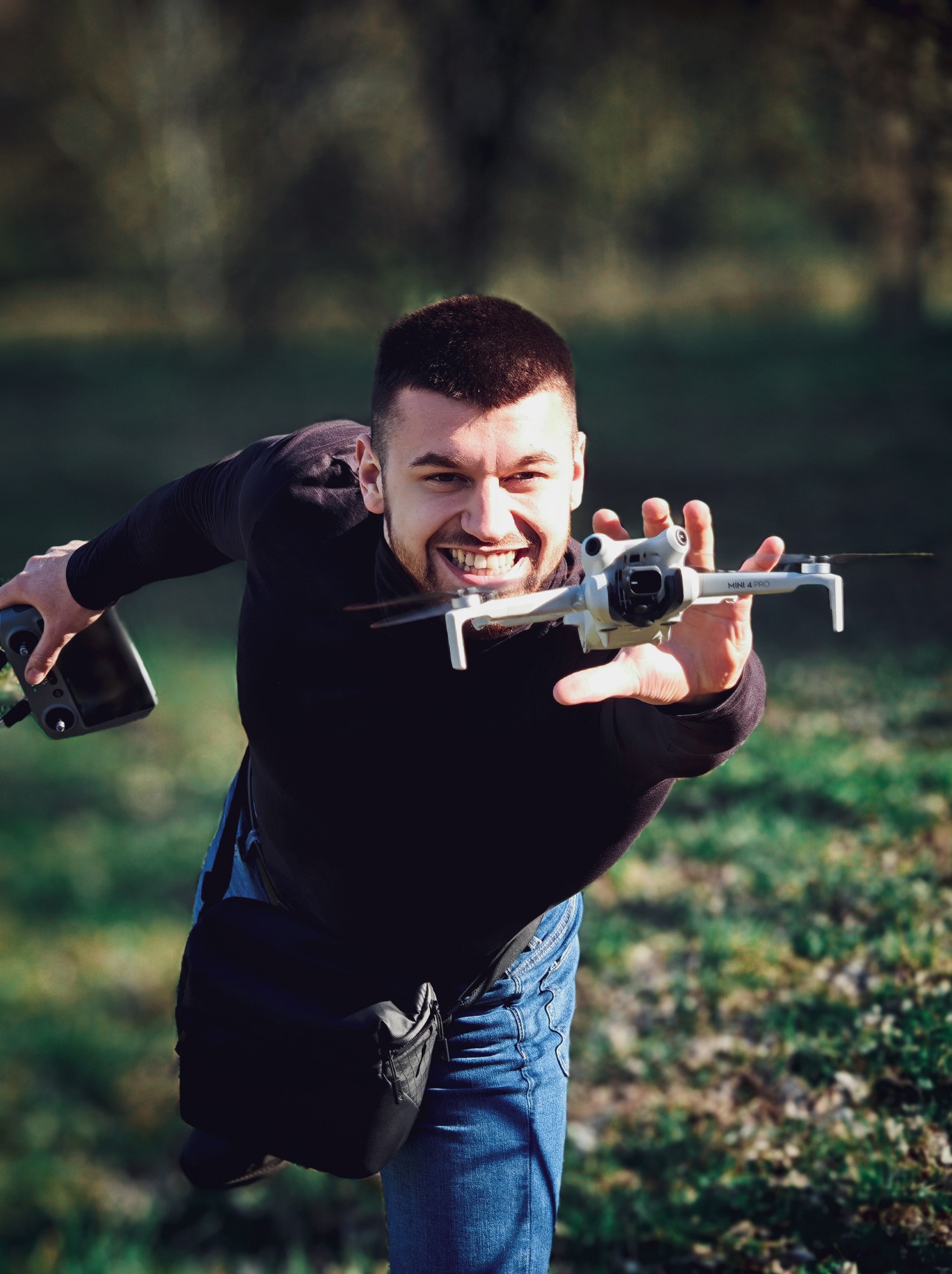 Uśmiechnięty mężczyzna trzyma drona DJI Mini 4 Pro i kontroler na tle zieleni. Dynamiczne ujęcie z dołu, ukazujące pasję do filmowania z powietrza.