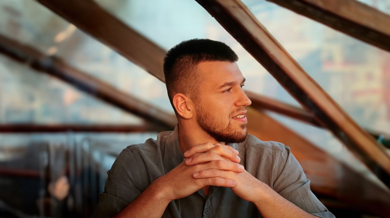 Młody mężczyzna z brodą, w szarej koszuli, spogląda w bok z zamyśleniem na tle rozmytej konstrukcji. Dłonie splecione przed sobą. Ujęcie z profilu.