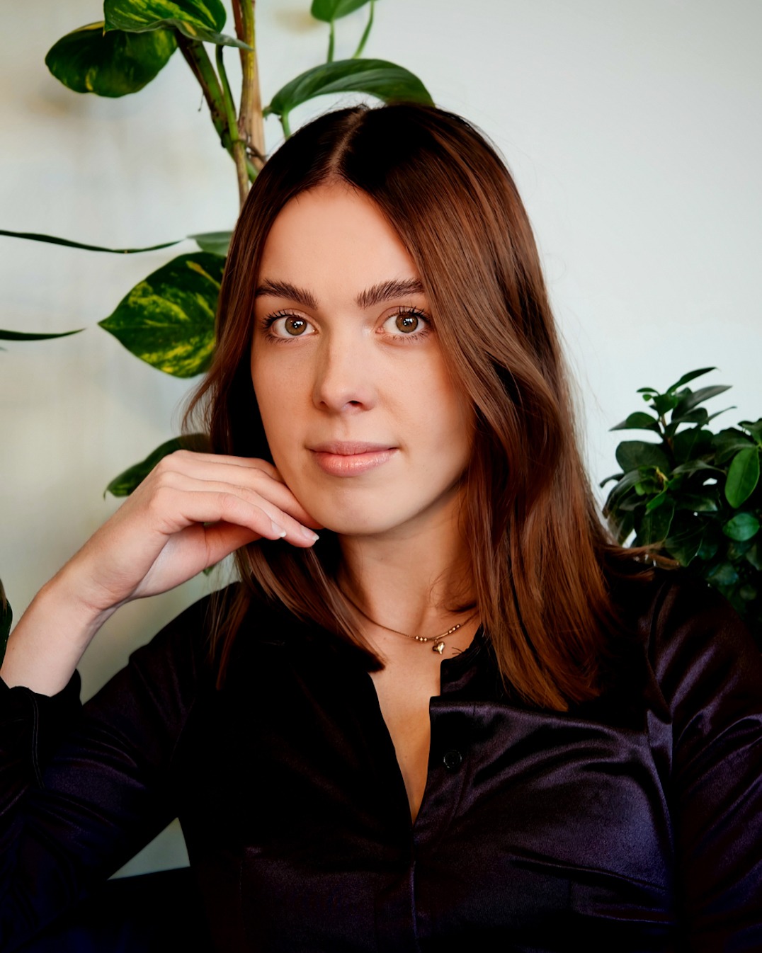 Portret kobiety z brązowymi włosami, w czarnej satynowej koszuli, z dłonią pod brodą, na tle zielonych roślin. Subtelny makijaż i złoty naszyjnik.