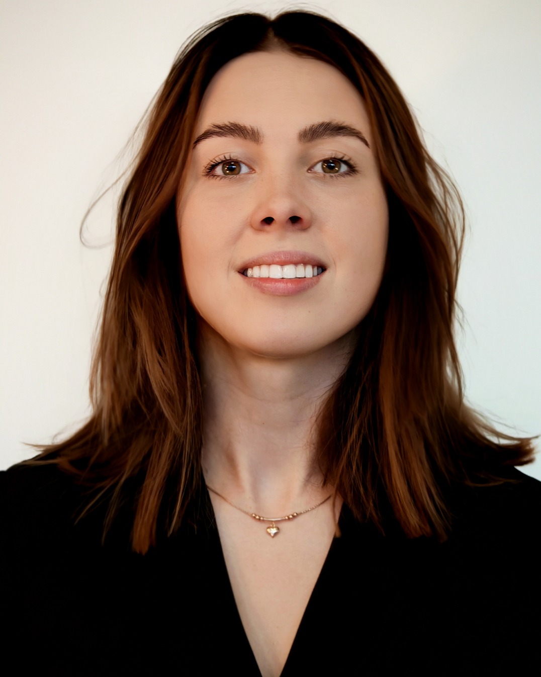 Portret uśmiechniętej brunetki z brązowymi oczami, w czarnej bluzce i delikatnym złotym naszyjniku na białym tle. Ujęcie centralne od klatki piersiowej w górę.