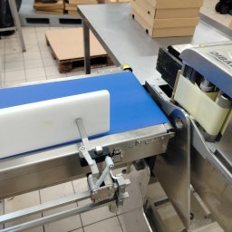 ART SYSTEM Rafał Lazar - Detali zdjęcia przedstawia automatyczną linię produkcyjną z niebieskim pasem transmisyjnym i urządzeniem etykietującym marki Dibal w tle, widoczne elementy konstrukcyjne ze stali nierdzewnej,...