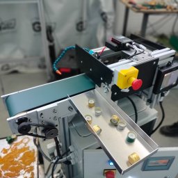 ART SYSTEM Rafał Lazar - Automatyczna linia produkcyjna z taśmociągiem sortująca metalowe nakrętki, widoczny panel sterowania i czujniki. W tle zamazane logo firmy.