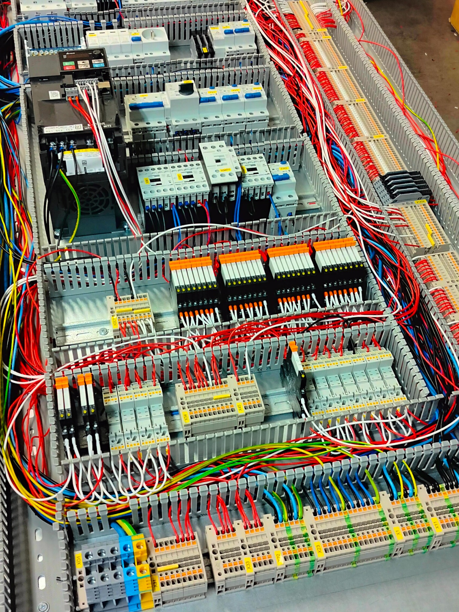Szafa sterownicza z bogatym okablowaniem, widoczne liczne moduły, bezpieczniki i złączki, przewody w kolorach czerwonym, białym, niebieskim, zielonym i żółtym starannie ułożone.