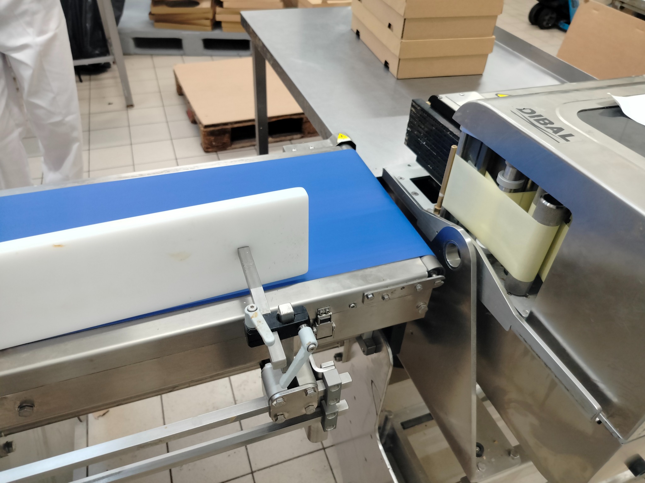 Detali zdjęcia przedstawia automatyczną linię produkcyjną z niebieskim pasem transmisyjnym i urządzeniem etykietującym marki Dibal w tle, widoczne elementy konstrukcyjne ze stali nierdzewnej,...