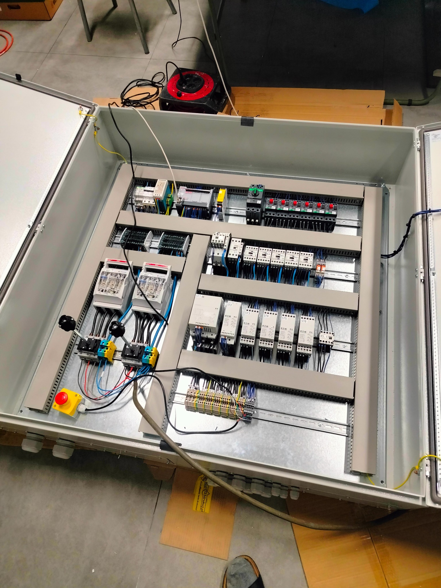 Otwarta szafa sterownicza z zainstalowanymi elementami elektrycznymi, widoczne przekaźniki, bezpieczniki, listwy zaciskowe i okablowanie, przycisk bezpieczeństwa na panelu, kabel zasilający...