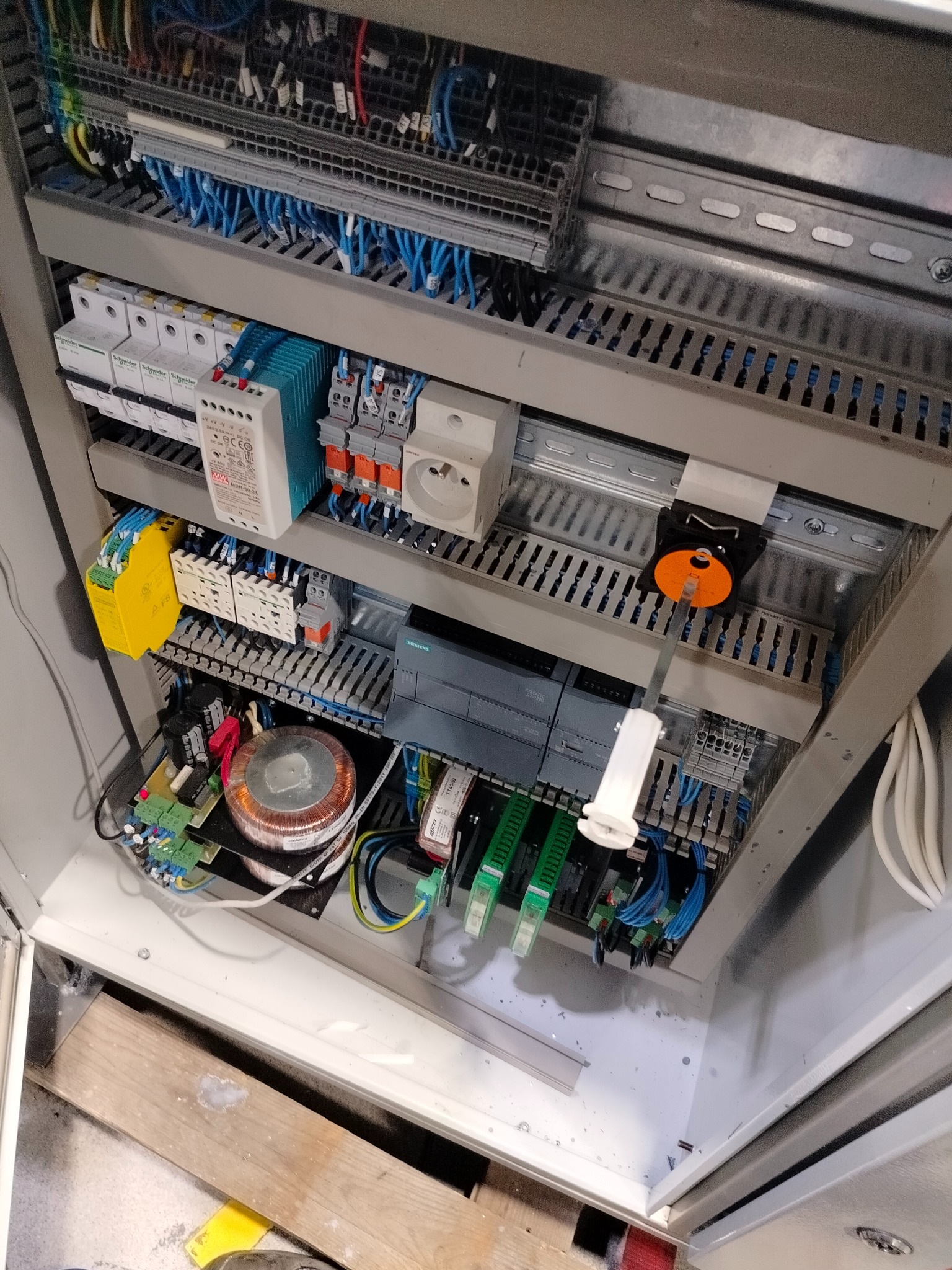 Wnętrze szafy sterowniczej z widocznymi elementami elektrycznymi: transformator, sterownik PLC Siemens, bezpieczniki, listwy zaciskowe i okablowanie. Widoczne przewody w kolorach niebieskim...